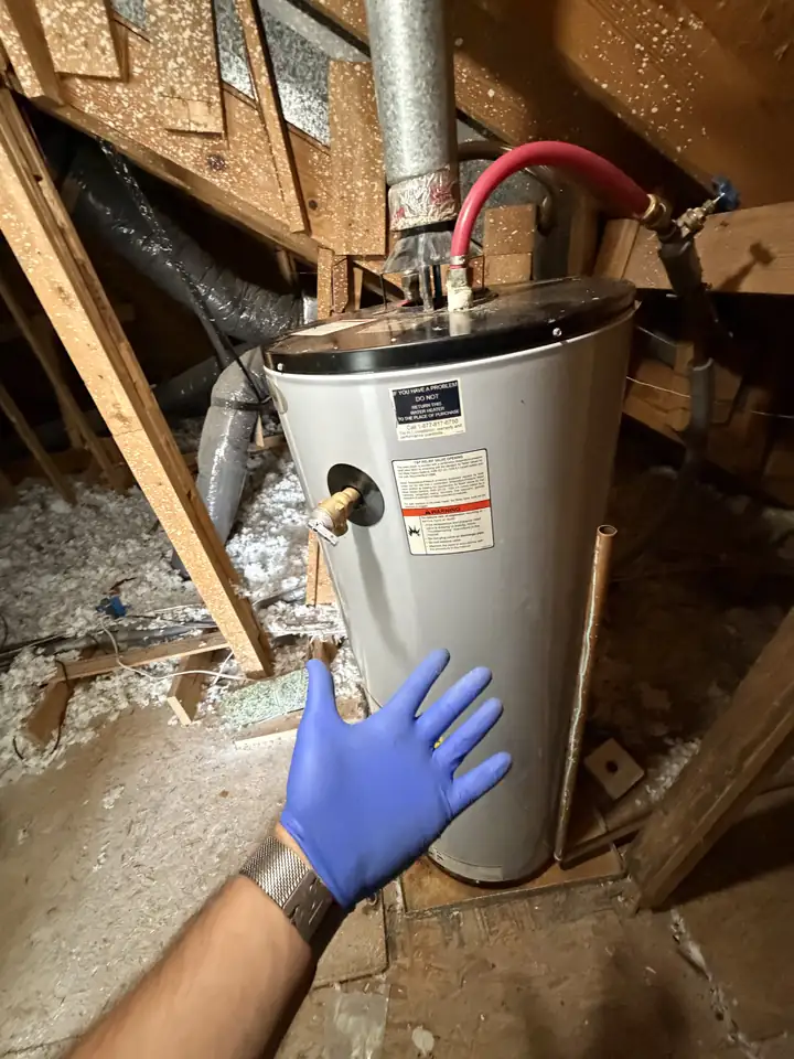 Old gas water heater in an attic, a bare T&P valve on its side and a red PEX line bent in a tight arc off the top, a gloved hand pointing, Allen