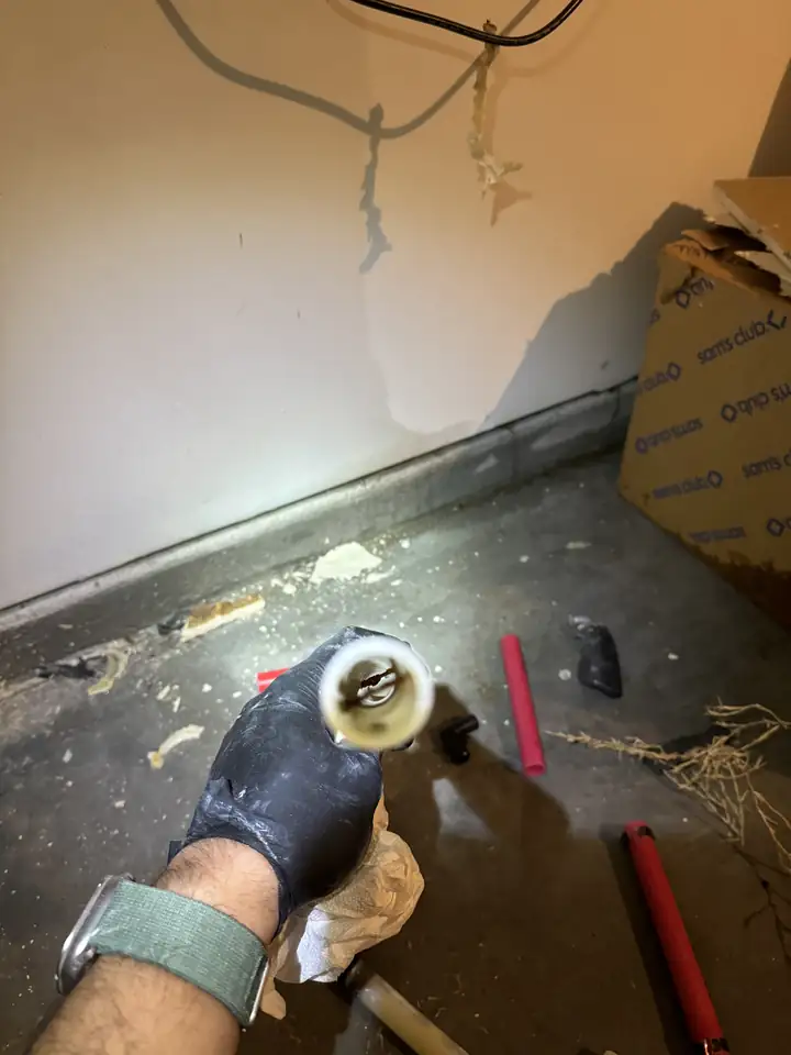 Gloved hand holding a cut piece of PEX pipe with a rusted nail inside it, water on the concrete floor, McKinney