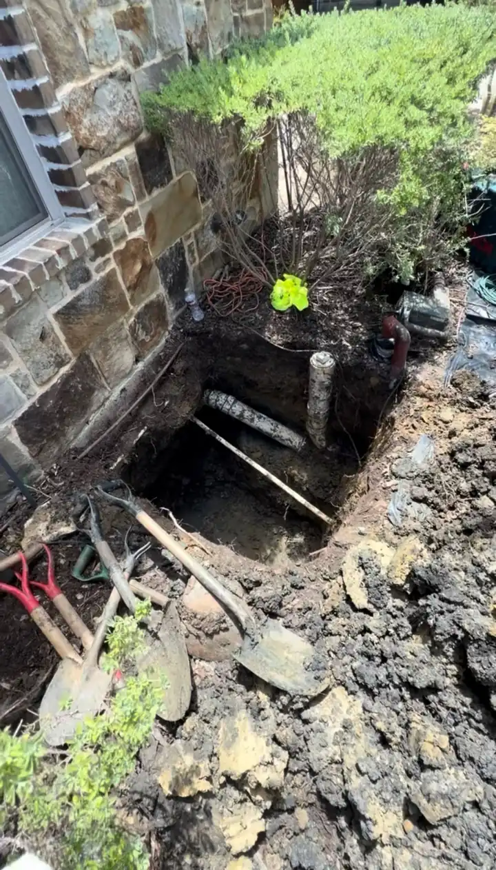 Hole dug next to a house foundation to reach a broken main sewer line, shovels beside it, Frisco