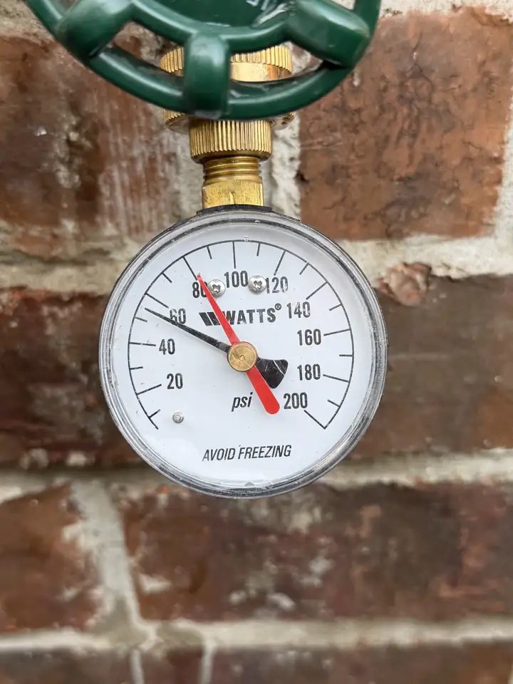 Water pressure gauge on an outside spigot at a brick wall, the black needle at about 56 PSI, McKinney