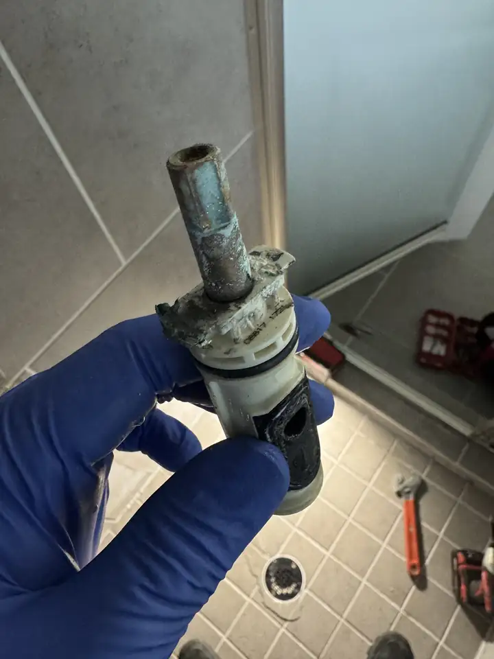 Corroded Moen shower cartridge with a green stem held in a gloved hand in a tiled shower, Prosper