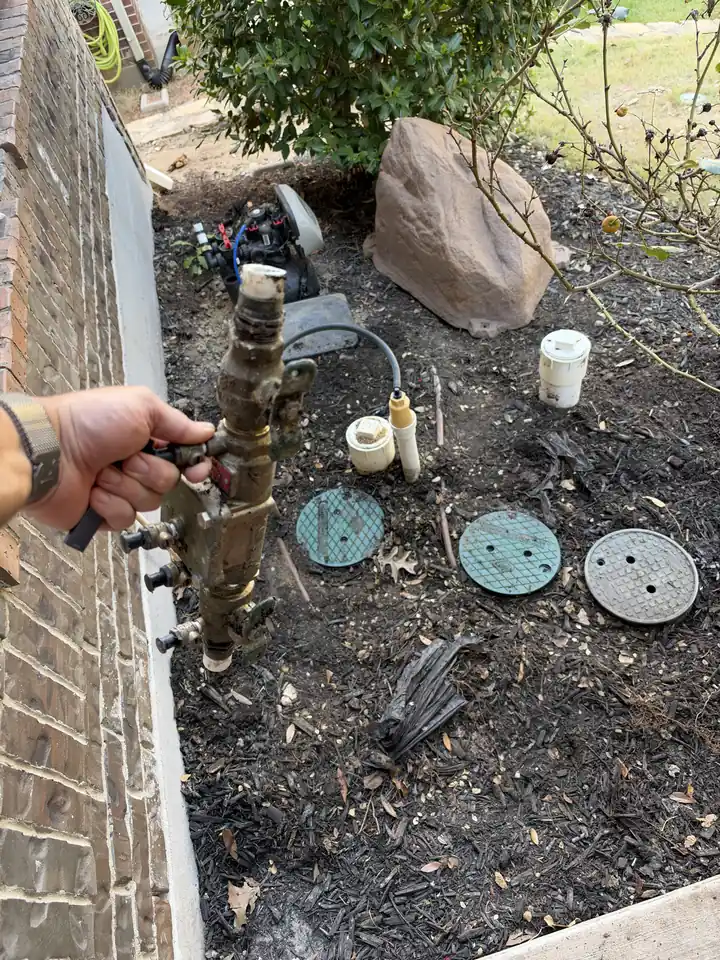 Hand holding a corroded valve assembly dug out of a flower bed, valve box lids and a decorative rock behind it, McKinney