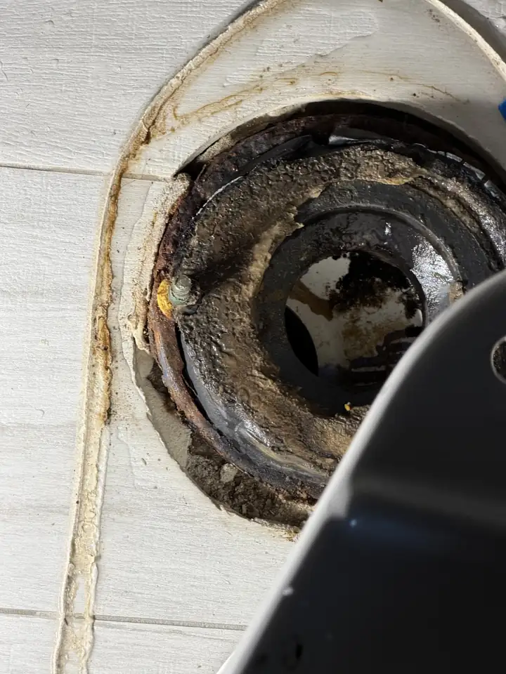 Rusted toilet flange with the old wax ring on a white tile floor, a scraper at the edge, Allen